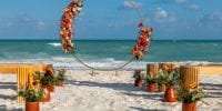 A floral arch and flower arrangements prepared for a destination wedding on a sandy beach by the ocean.