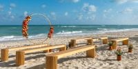 Wooden benches and a floral arch arranged for a destination wedding ceremony on a sandy beach by the ocean.