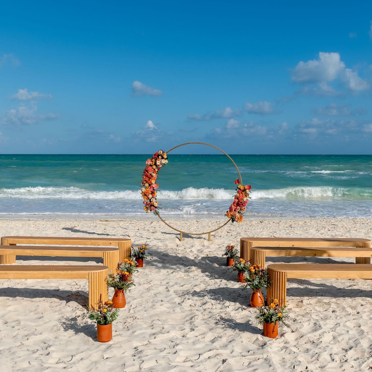 Wooden benches and a floral arch arranged for a destination wedding on a sandy beach by the ocean.