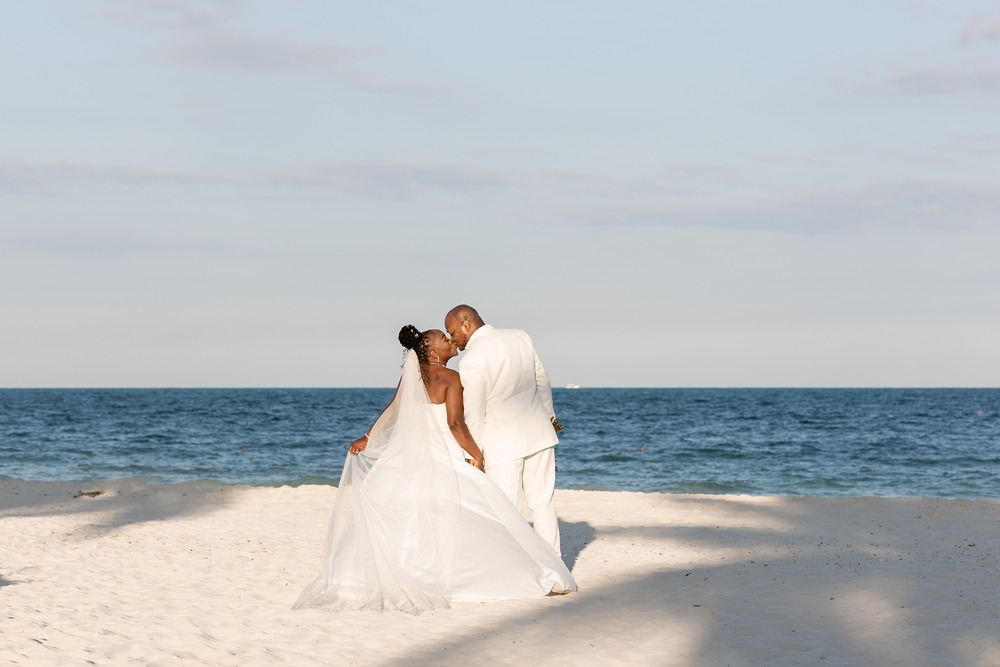 Catherine and Carlos stand on a sandy beach in white wedding attire, gazing at the ocean.