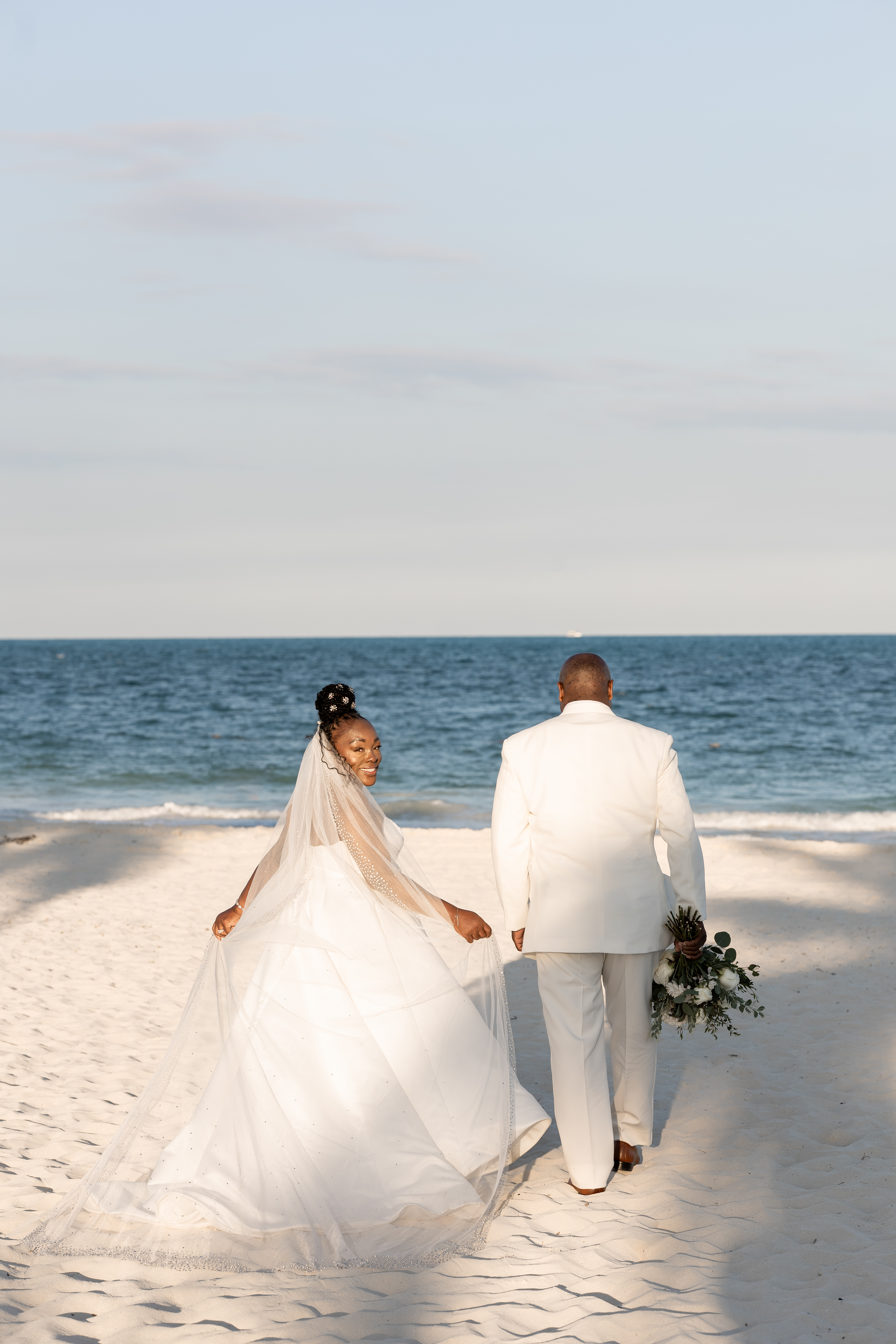 Catherine and Carlos (Copy), in white attire, walk hand in hand on a beach at their destination wedding.