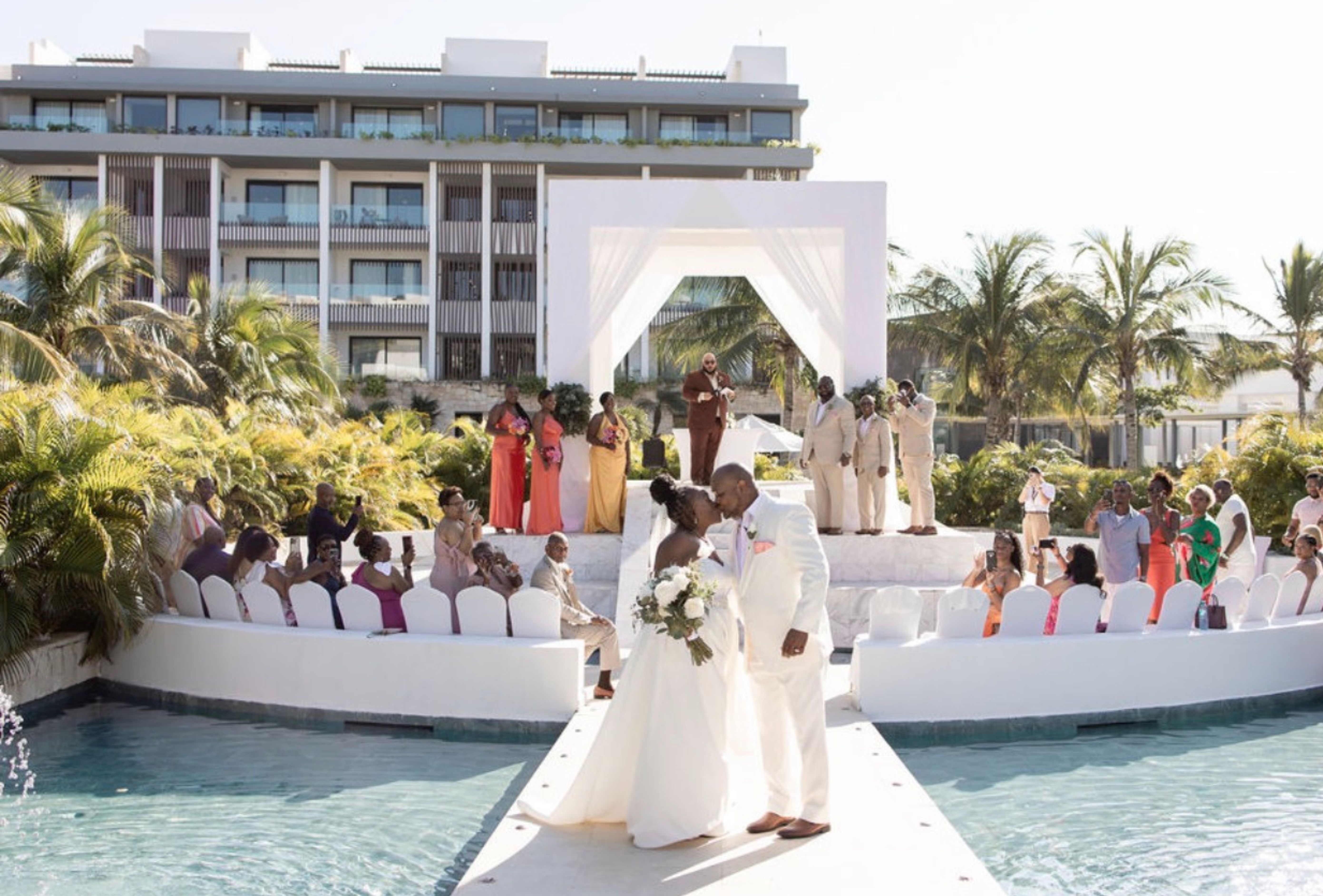 Catherine and Carlos share a kiss outdoors at their destination wedding, guests seated close by.