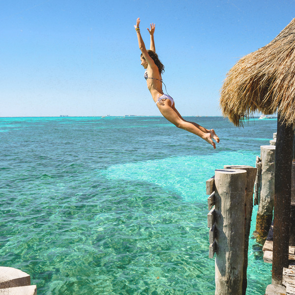 Woman in a bikini leaps off a wooden dock into turquoise ocean water at a tropical destination wedding.