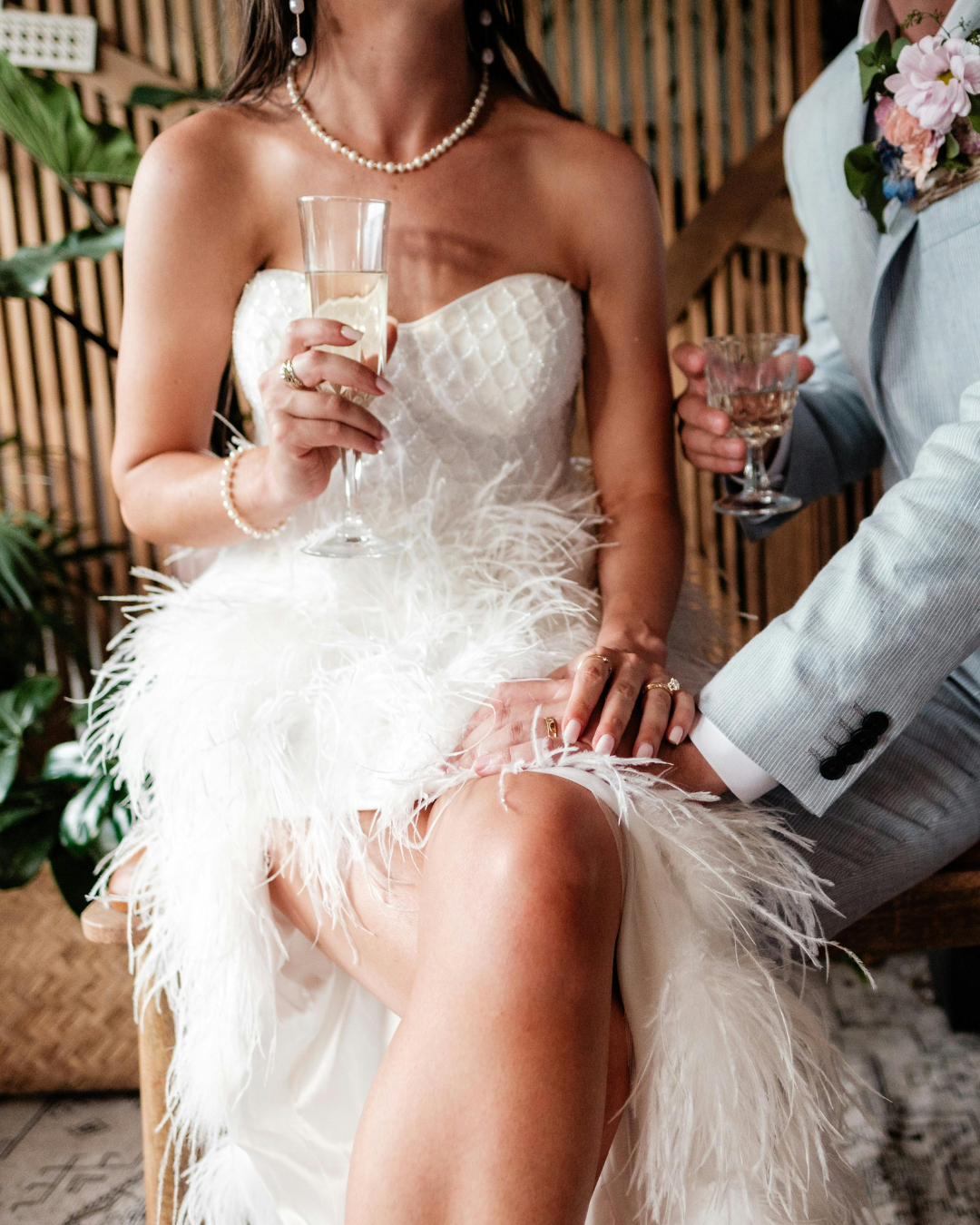 Bride in a feathered white gown holding champagne beside her partner at a stylish destination wedding.