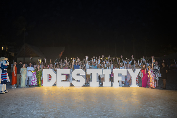 A group celebrates a destination wedding on sandy ground at night behind large "DESTIFY" letters, arms up.