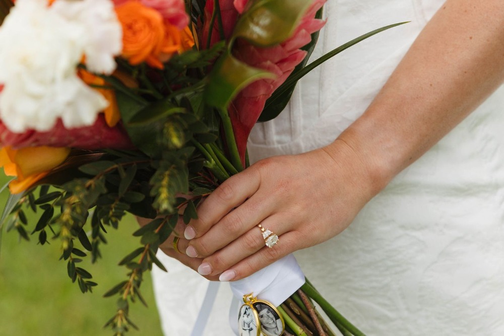 At a destination wedding, someone in white holds a bouquet and displays an engagement ring on their left hand.