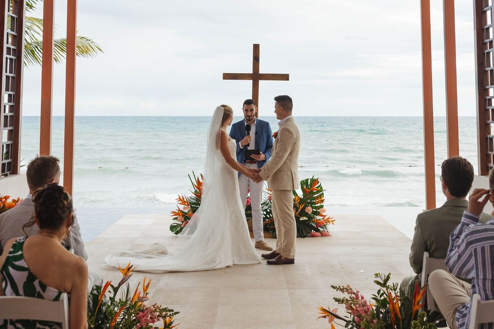 Hannah and Ben stand with an officiant during their destination wedding at an indoor beachside venue.