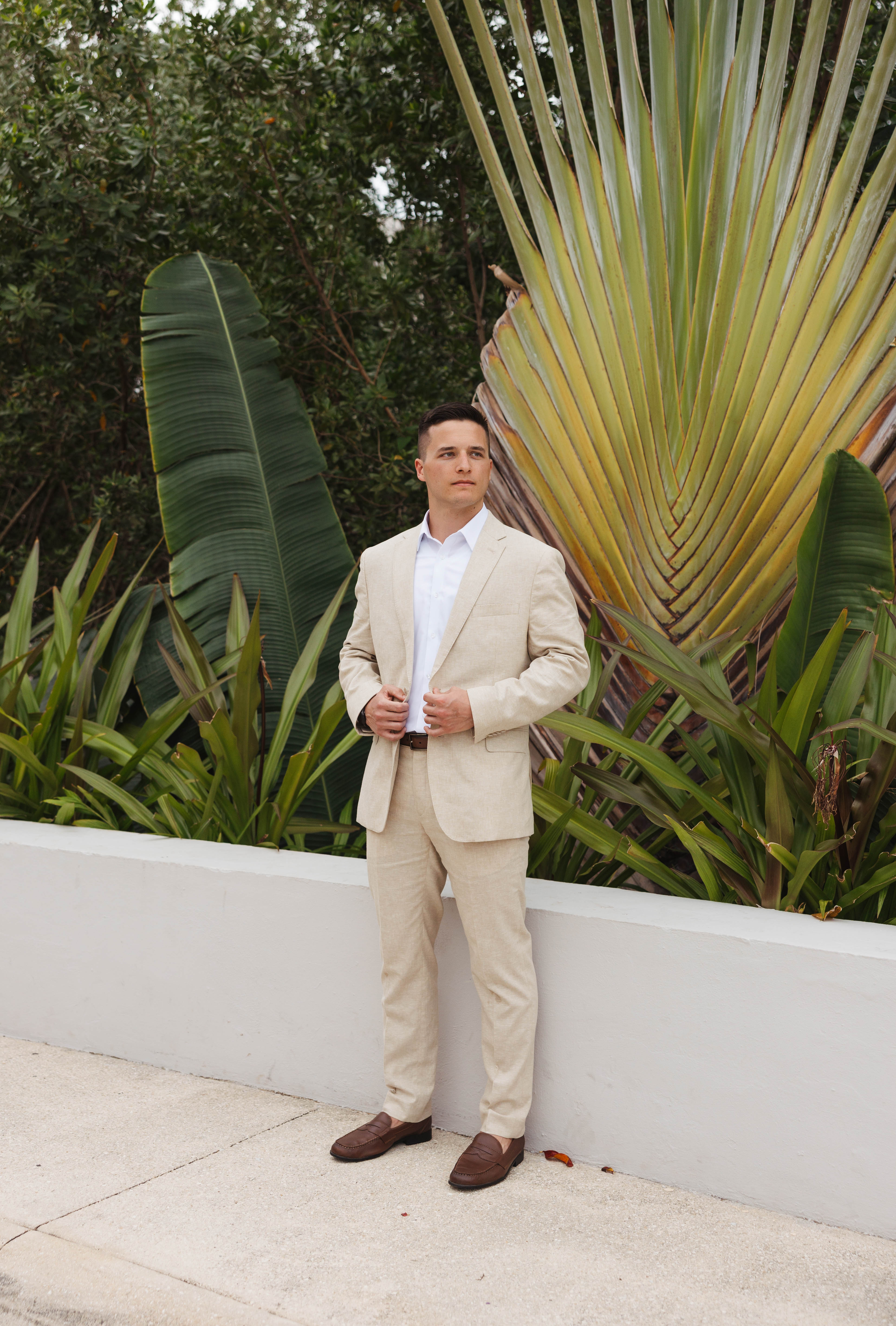 Man in a beige Tiffany and Anthony suit poses by tropical plants and a white wall at a destination wedding.