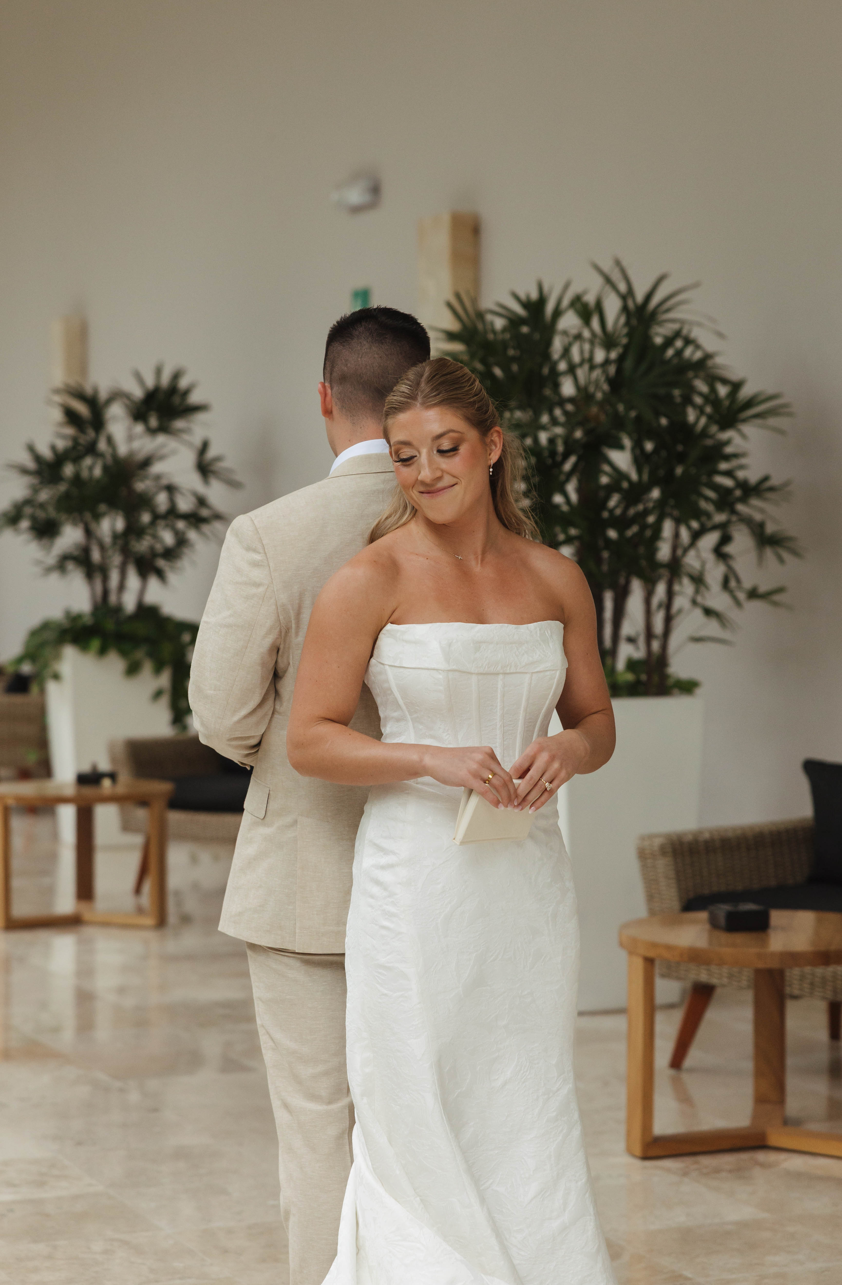 Tiffany and Anthony stand back-to-back indoors; Tiffany smiles, holding an envelope for their wedding.