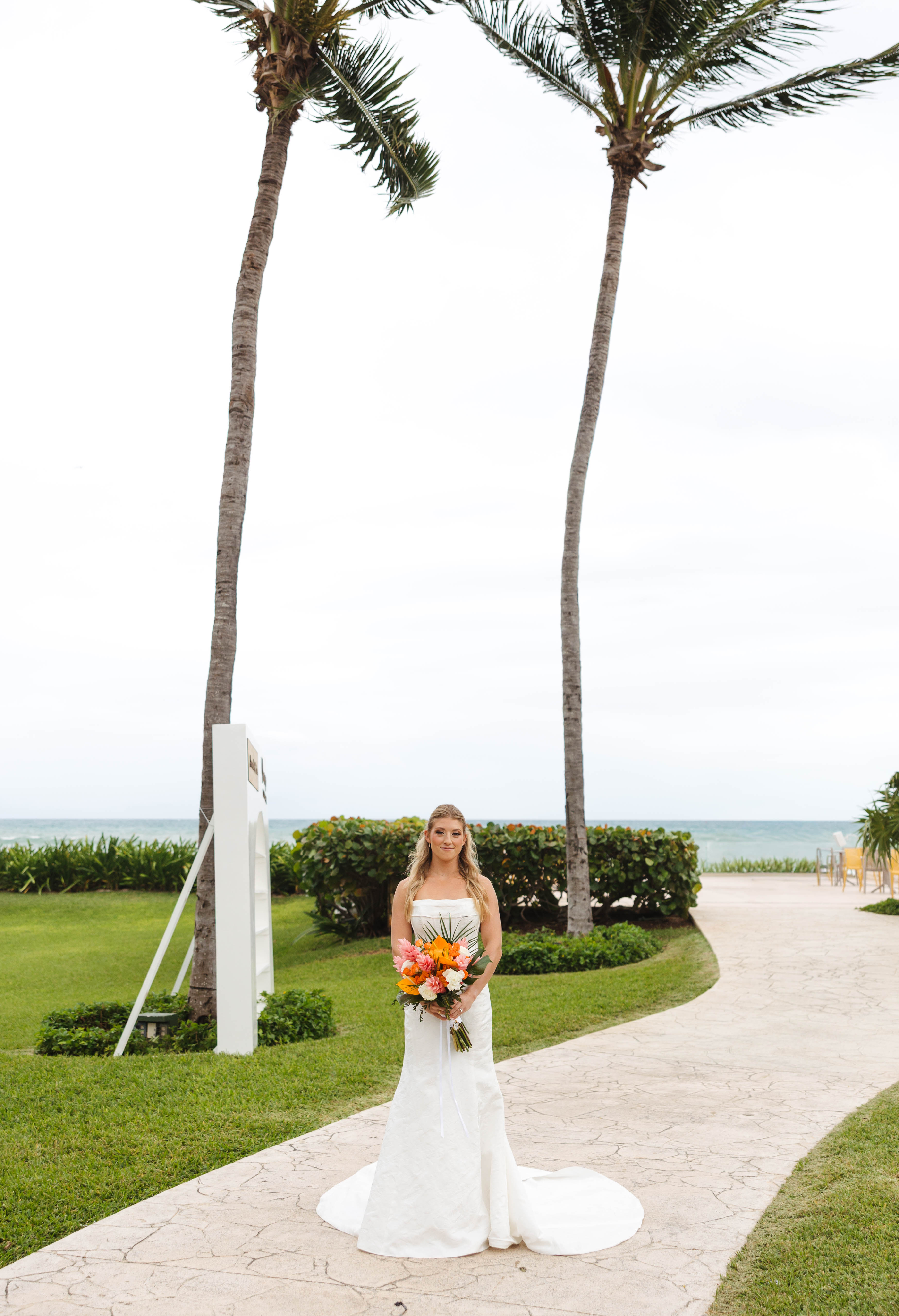 Bride in white dress stands on a palm-lined path by the ocean at a beautiful destination wedding.