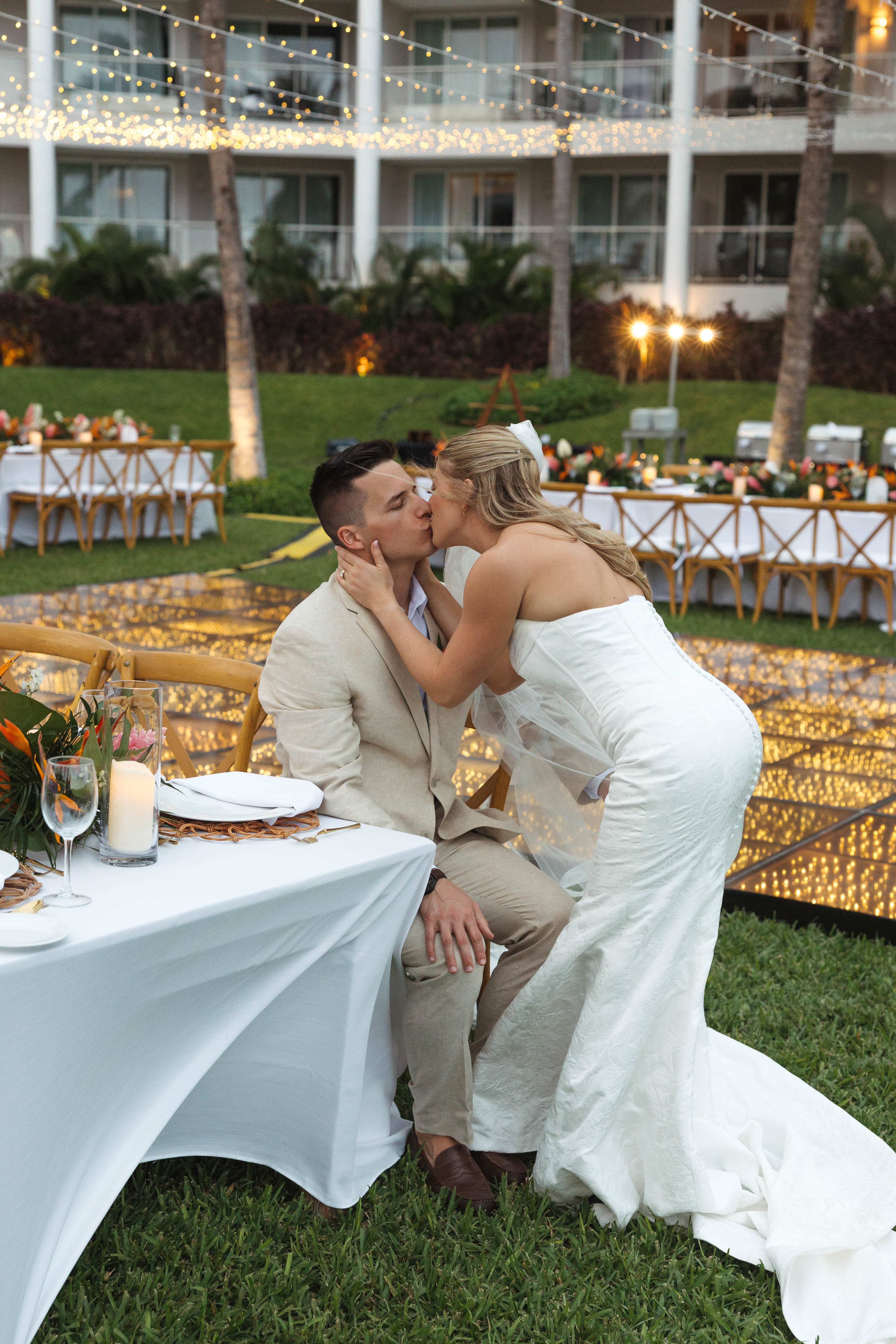 Tiffany kisses Anthony on the cheek at a destination wedding reception under glowing string lights.