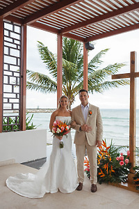 Tiffany and Anthony pose indoors by the ocean at their destination wedding, holding tropical flowers.