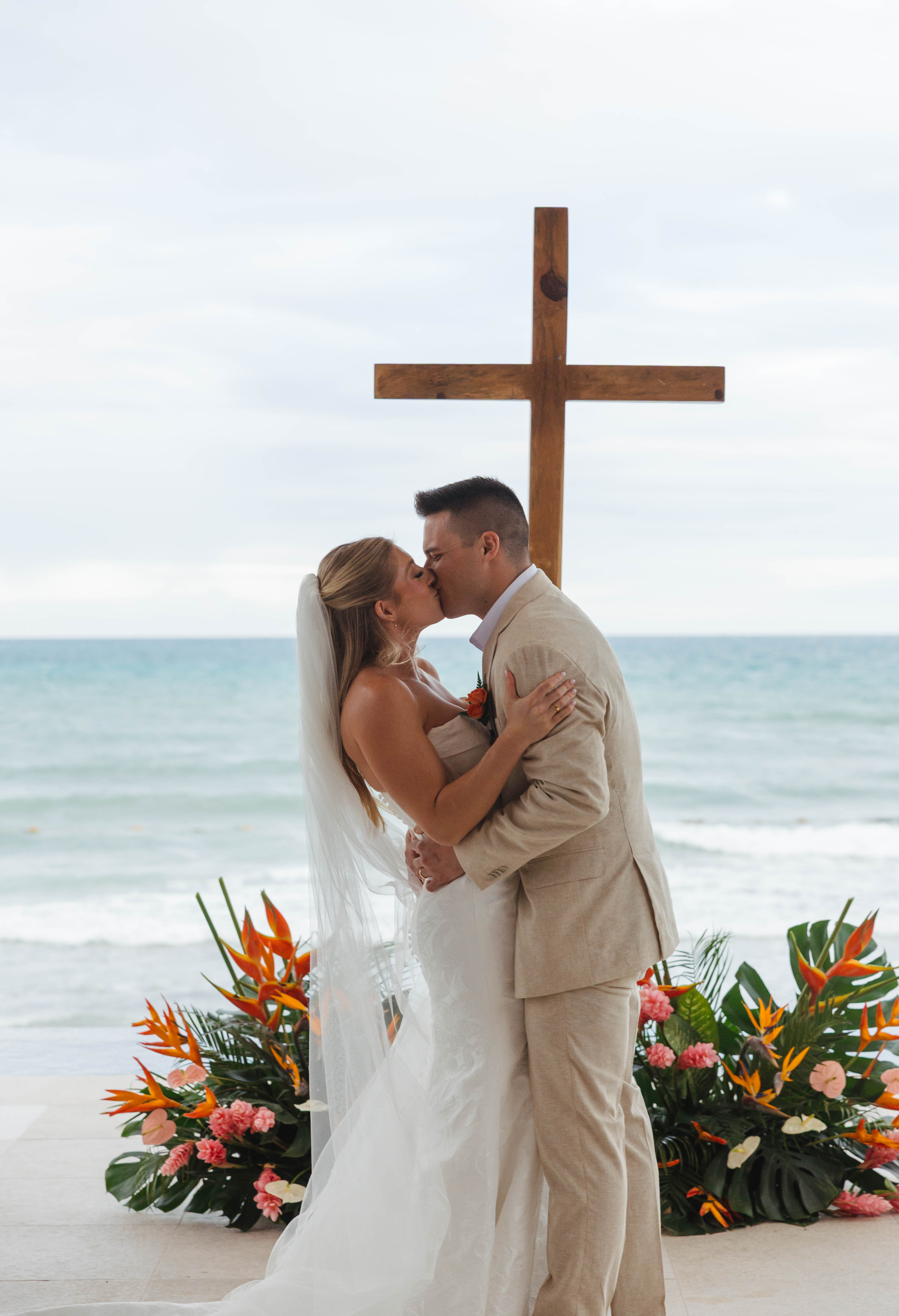 Tiffany and Anthony share a kiss by the ocean at their destination wedding, near a cross and flowers.