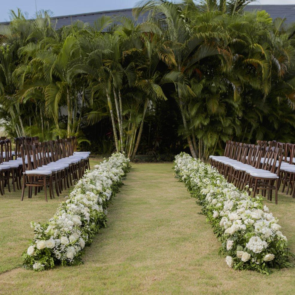 Destination wedding aisle adorned with white flowers, wooden chairs, and palm trees in the background.