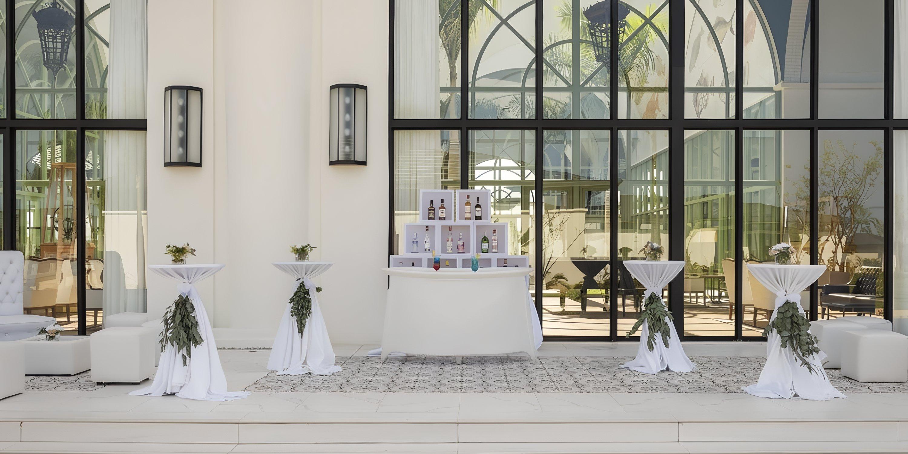 Wedding outdoor bar with cocktail tables in white linens and greenery, set before large windows.