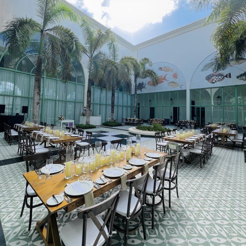 Long wooden tables set for a destination wedding in a courtyard with palm trees and patterned tile floor.