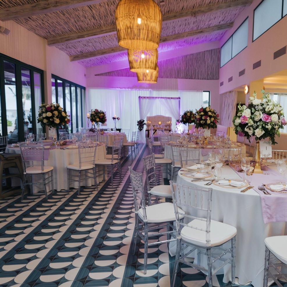 Elegant destination wedding reception indoors featuring round tables, floral centerpieces, and clear chairs.