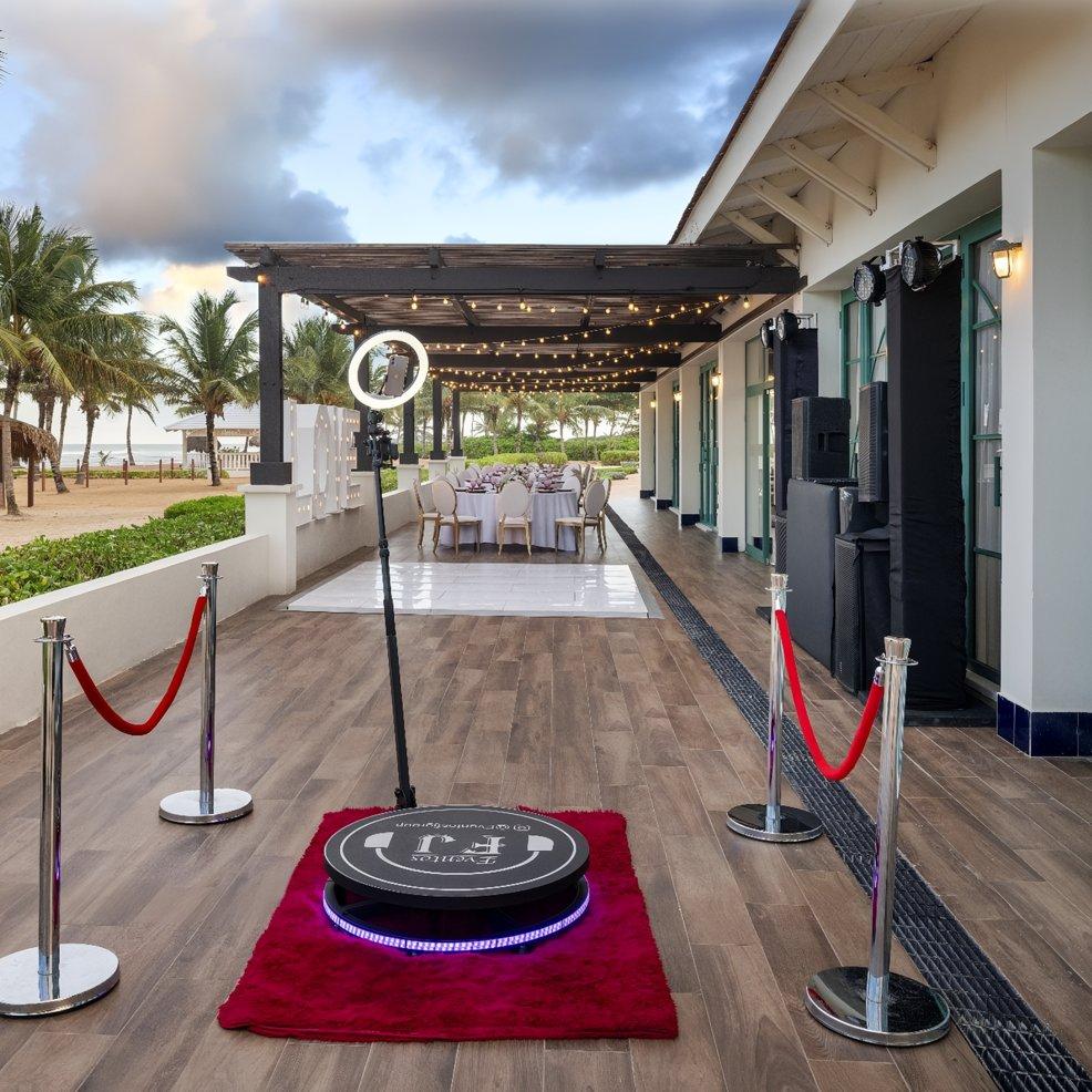 Destination wedding outdoor setup featuring a photo booth, red carpet, and dining area beneath string lights.