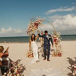 A couple walks hand-in-hand beneath a floral arch on the sand during their destination wedding ceremony.