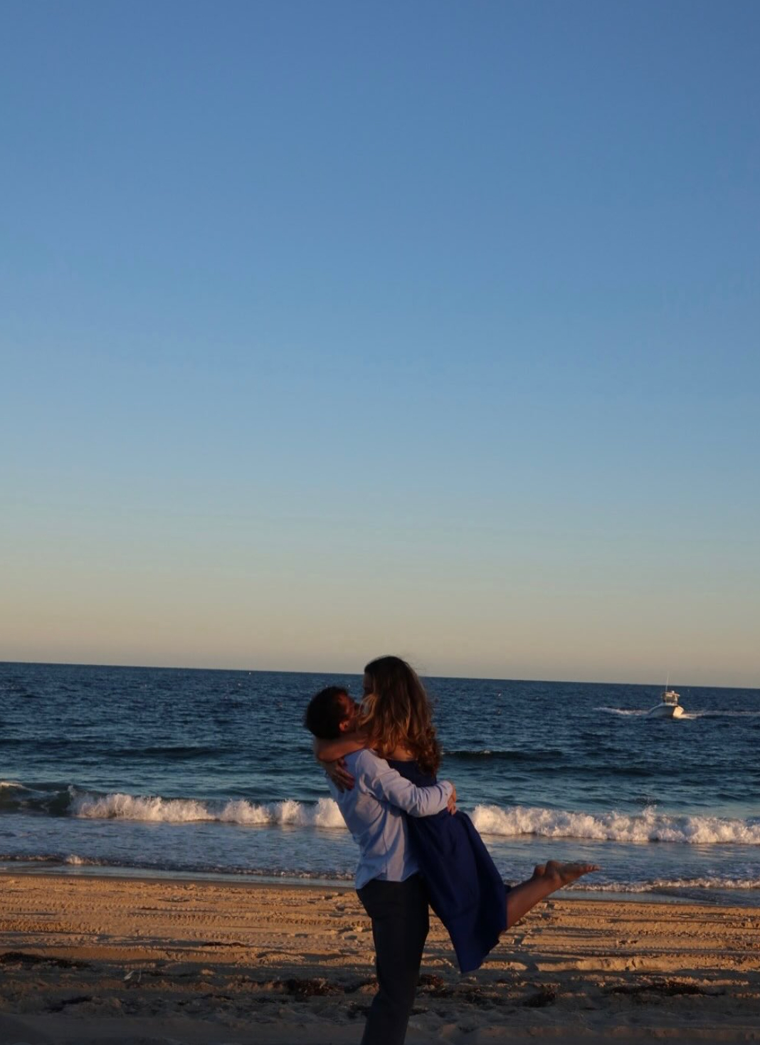 Couple shares a kiss during a destination wedding on a sandy beach, ocean and boat in the background.