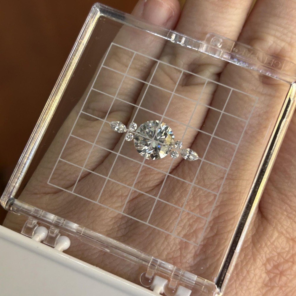 A wedding diamond ring with side stones displayed in a clear plastic grid box, held above a hand.