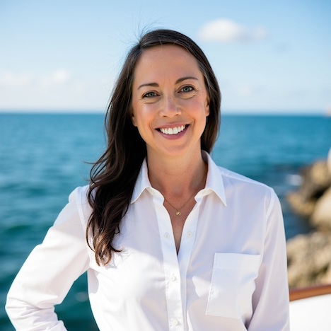 Woman in a white shirt standing outdoors at a destination wedding, ocean and blue sky in the background.