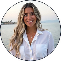 Woman with long wavy hair in a white shirt by water at sunset, smiling during a destination wedding.