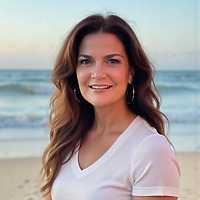 Woman with long brown hair in a white shirt, smiling on a beach at sunset during a destination wedding.