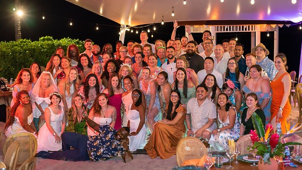A large group poses together at a destination wedding, gathered outdoors beneath glowing string lights.