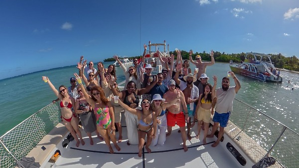 A group celebrating a destination wedding on a boat, raising their arms and smiling by the sunny shore.