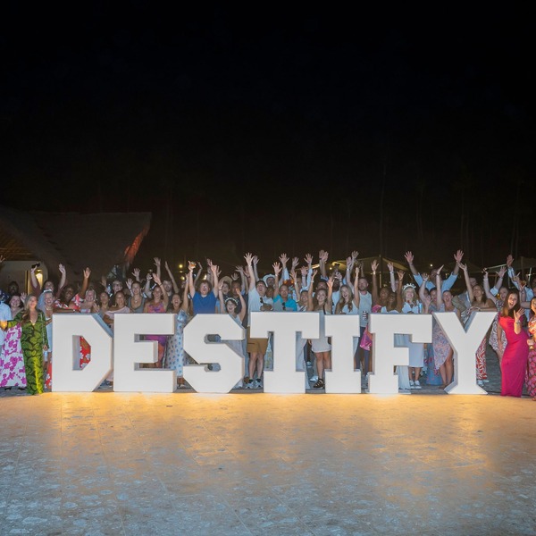 A large group celebrates a destination wedding at night behind giant &ldquo;DESTIFY&rdquo; letters, hands raised.