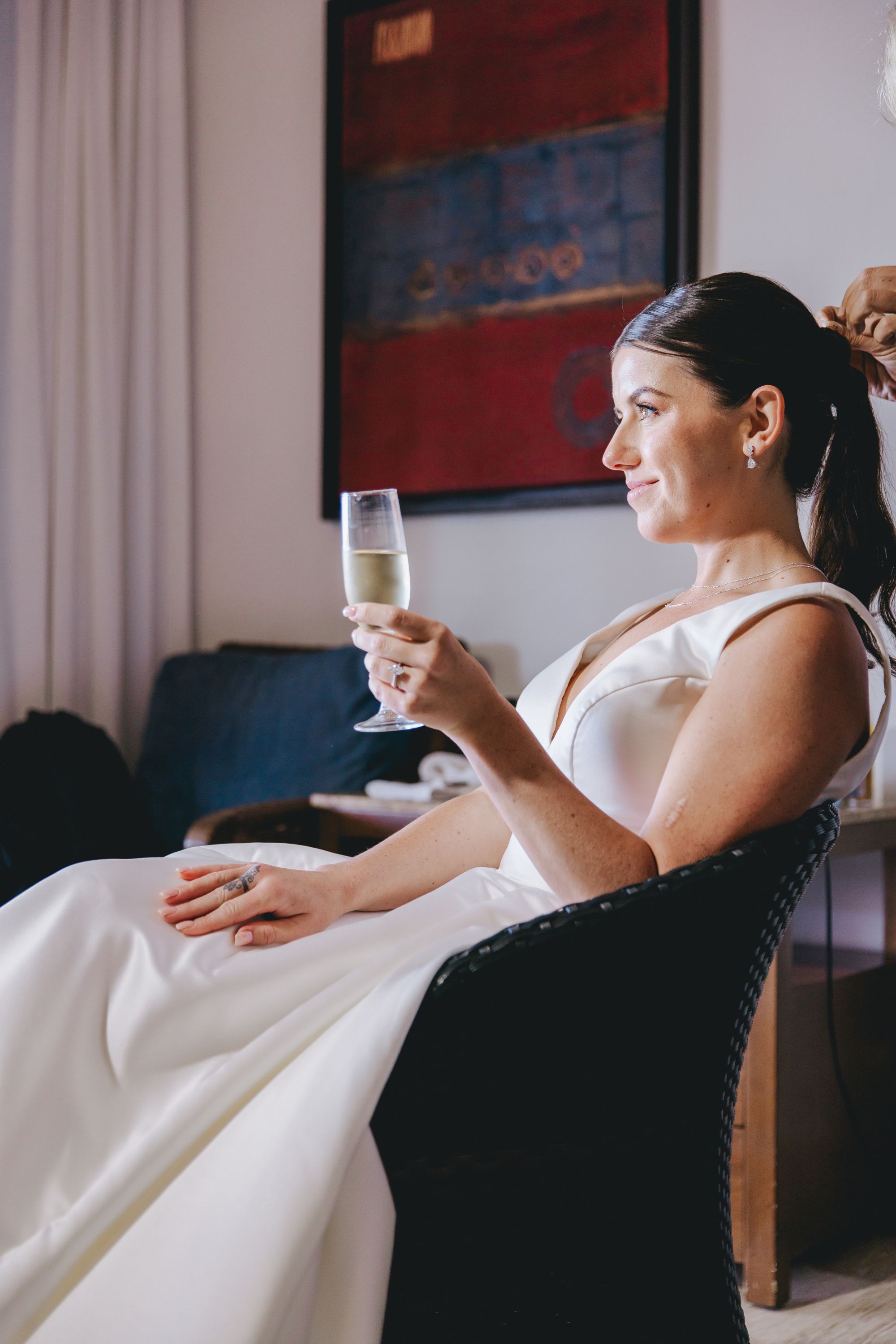 Woman in a Kathryn and Mike (Copy) white dress sits in a chair with champagne, facing right at wedding.