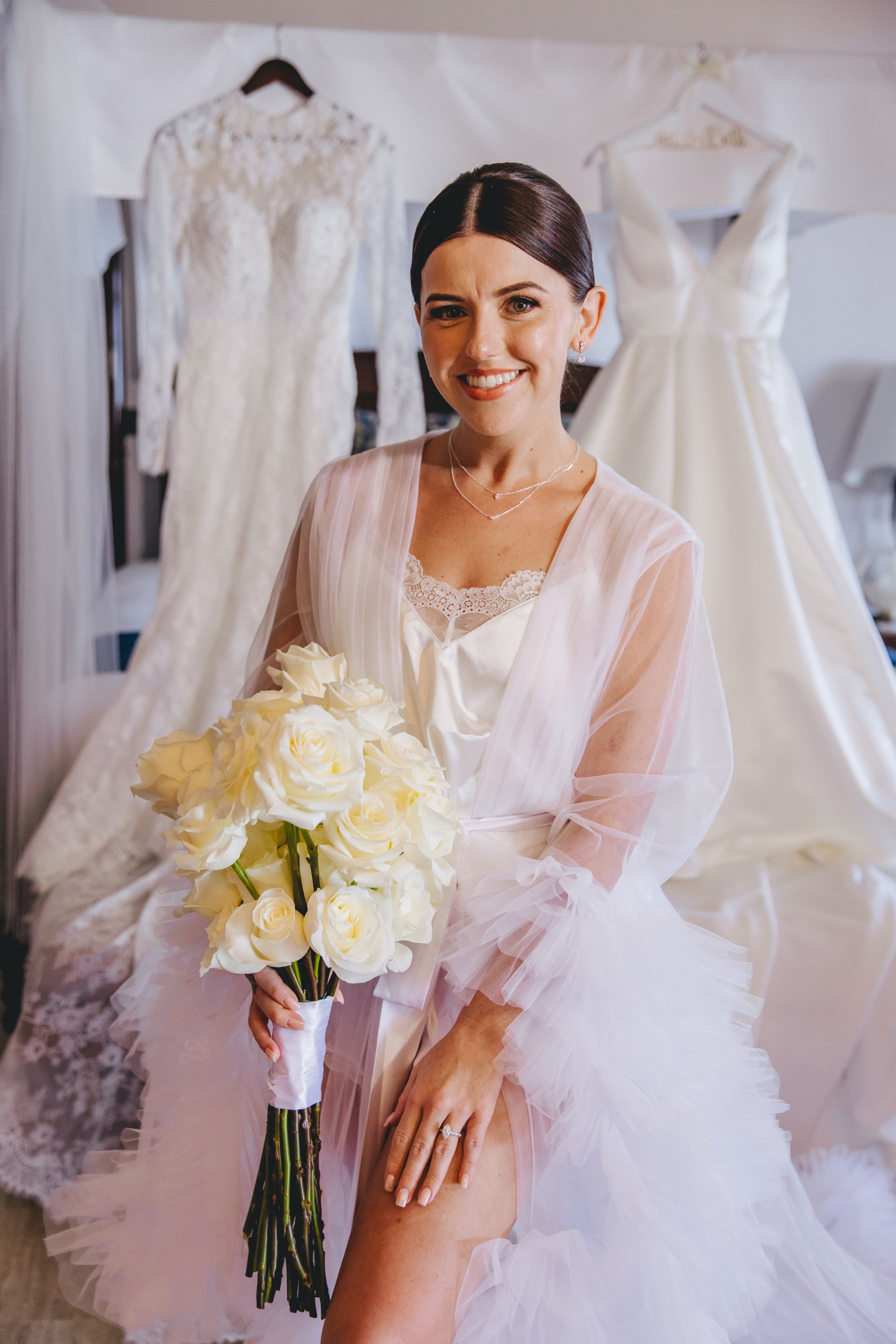 Bride in "Kathryn and Mike (Copy)" robe holds white roses near two wedding dresses at destination wedding.
