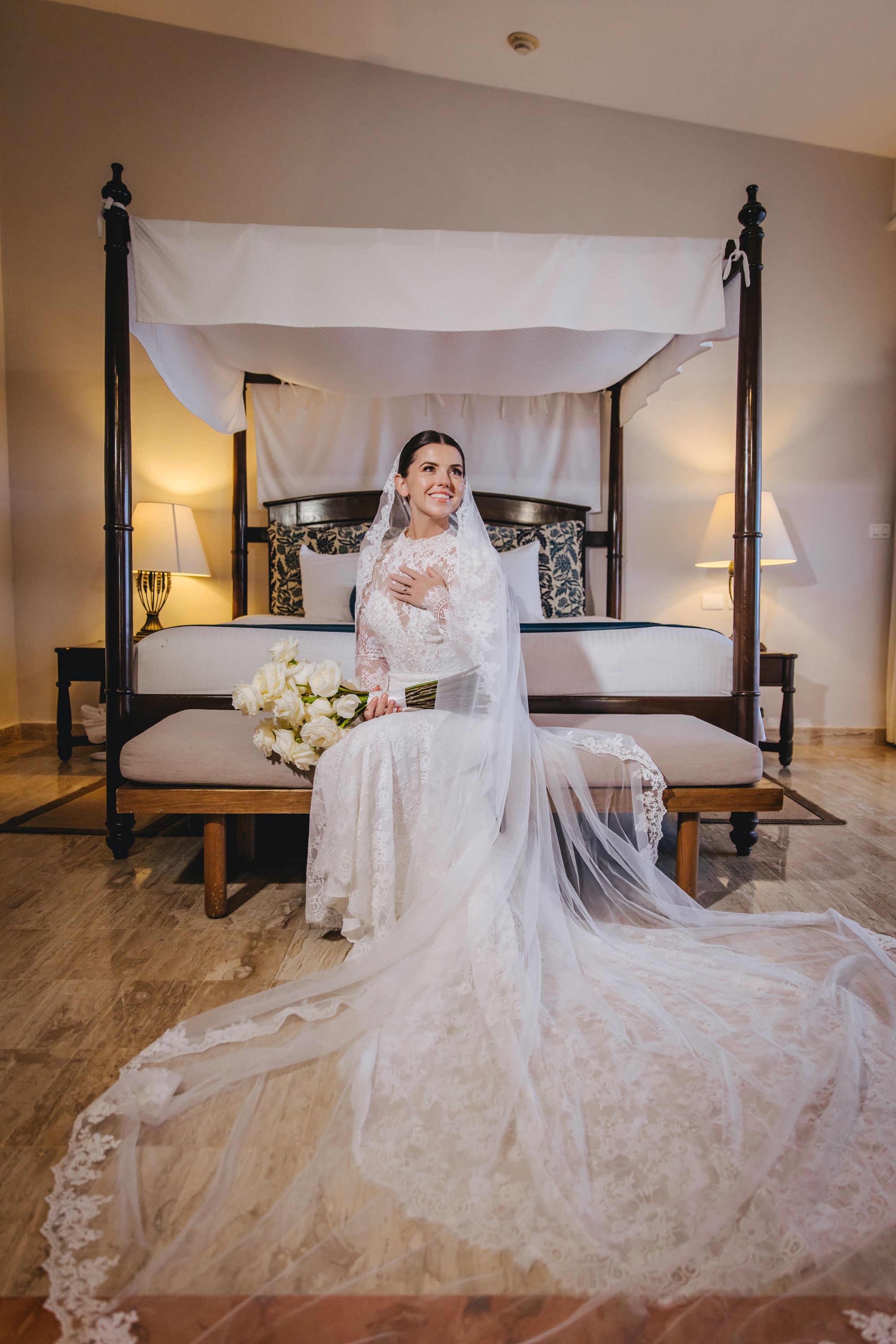 Bride in a lace gown with bouquet and long veil sits on a bench at her destination wedding.