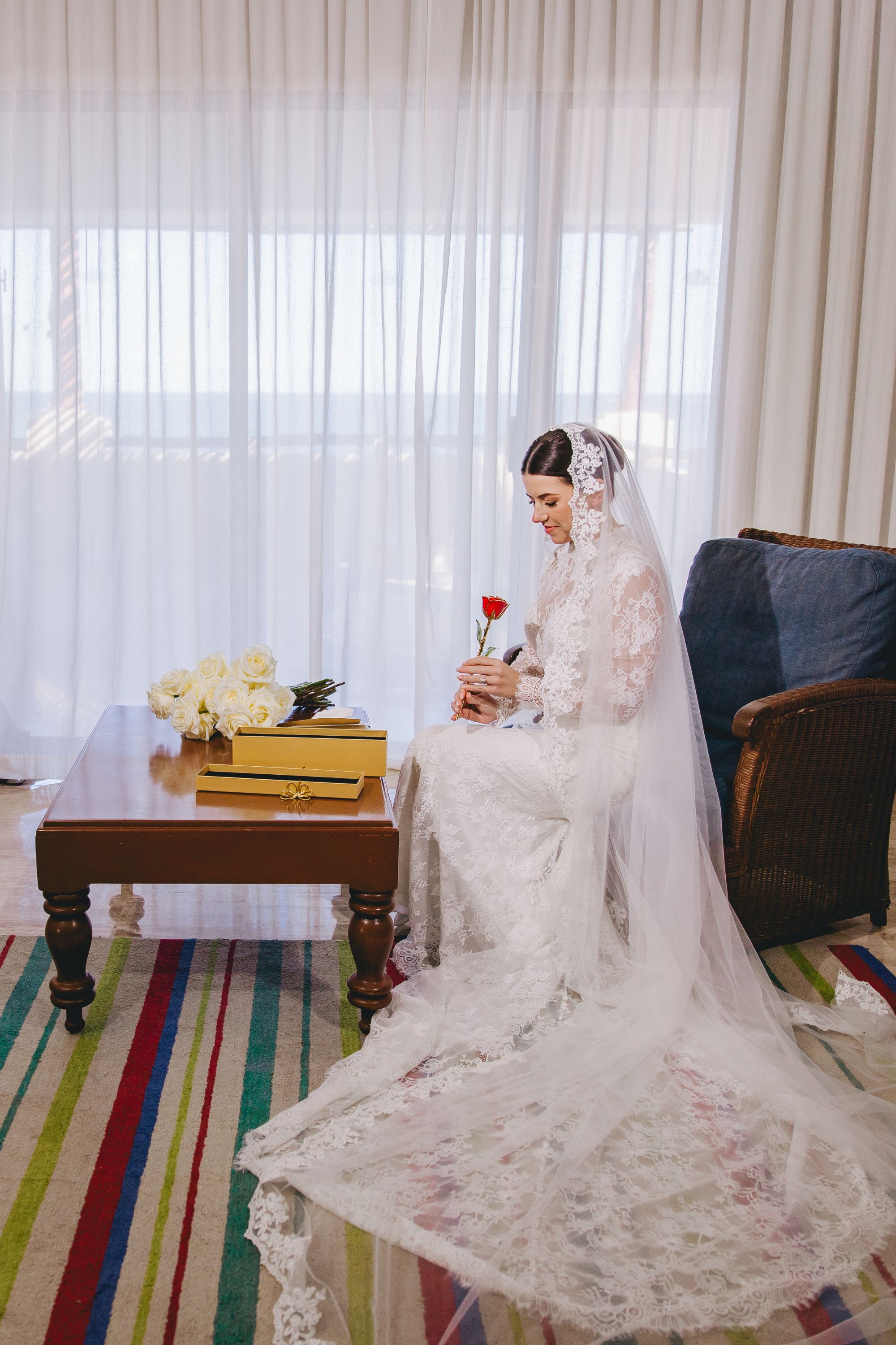 Bride in lace gown sits by flowered table at destination wedding, holding a single red rose.