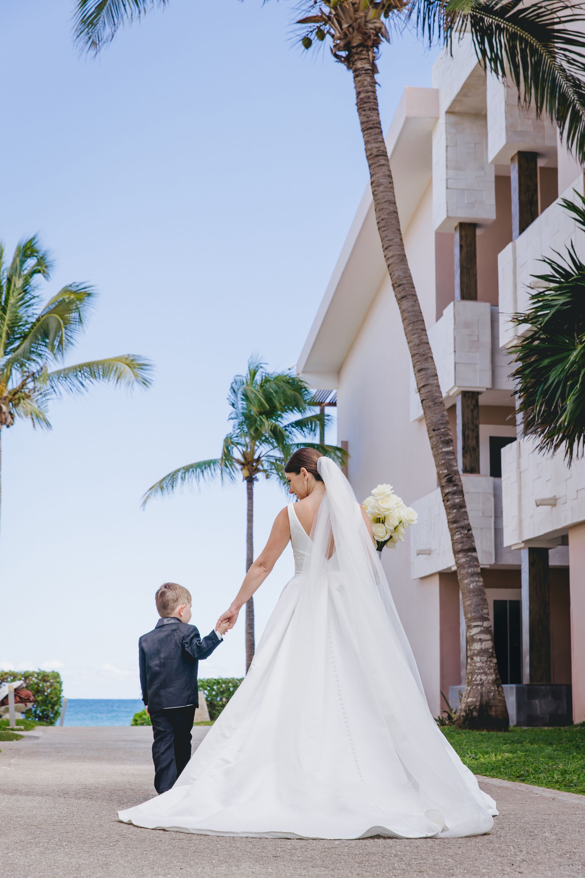 Bride in white gown with bouquet holds young boy's hand by palm trees at a destination wedding.
