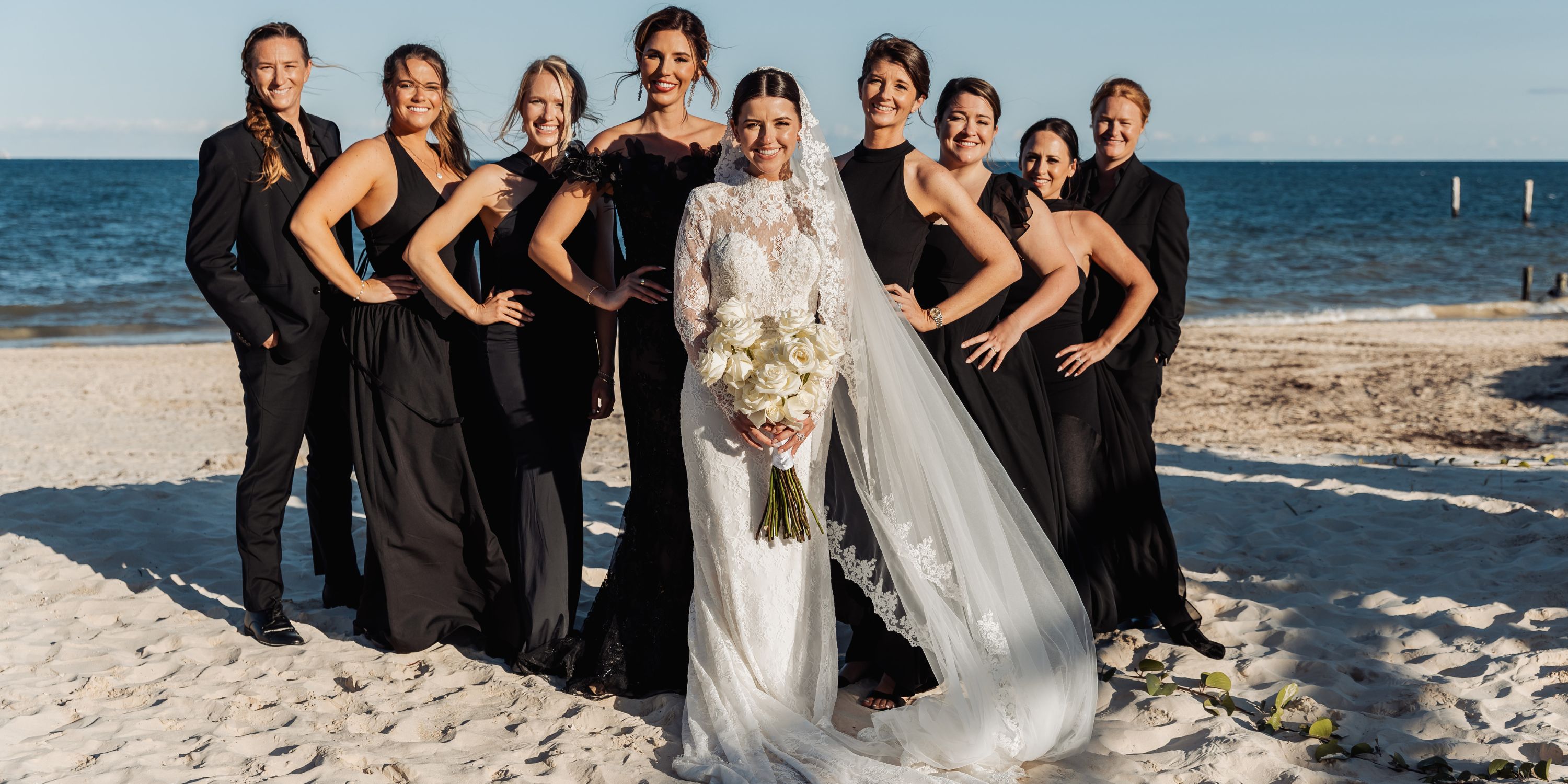 Bride in white holding a bouquet poses with bridesmaids in black at a beach destination wedding.