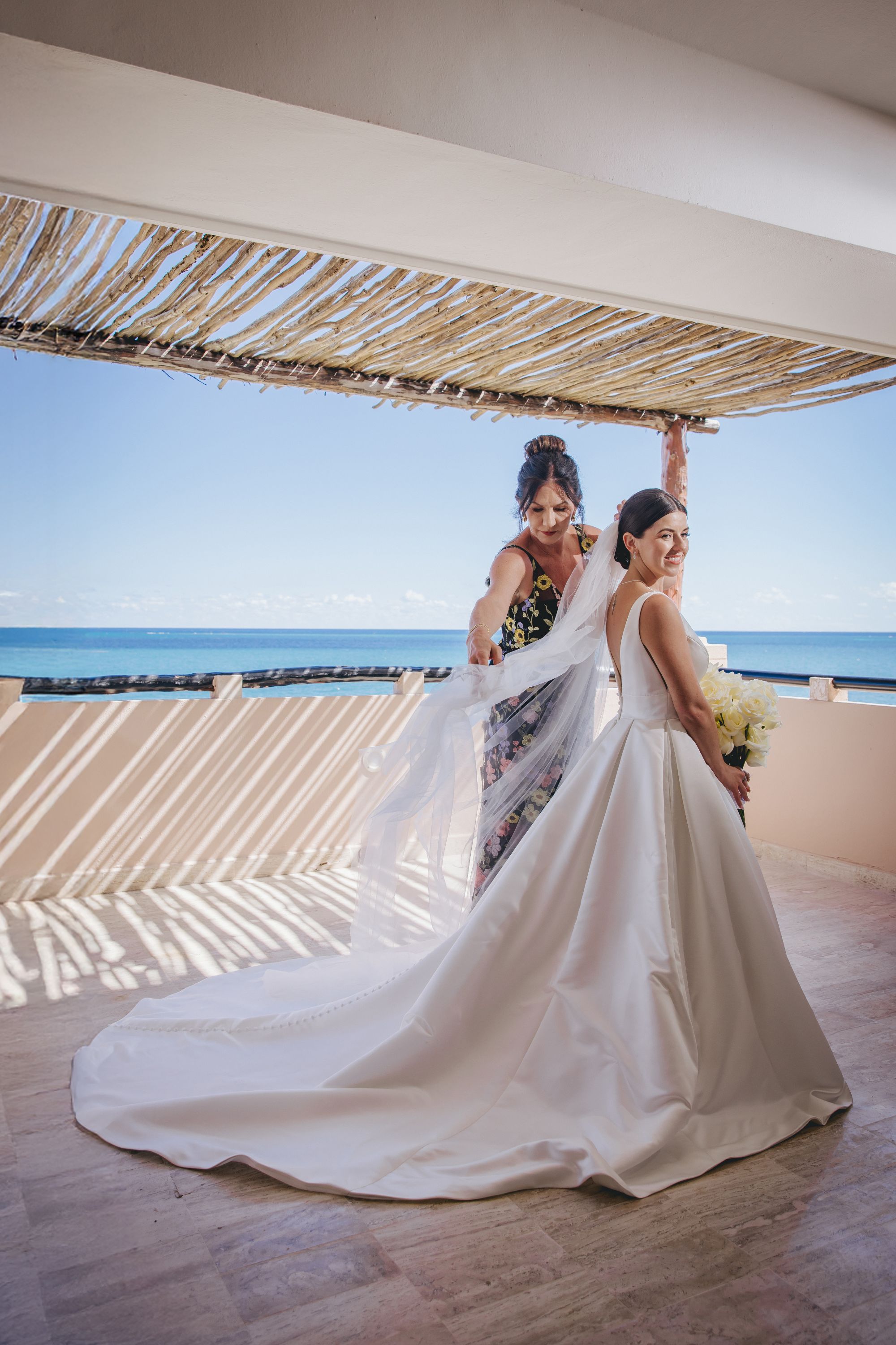Kathryn, in a white wedding gown, smiles as her veil is adjusted on a destination wedding balcony.