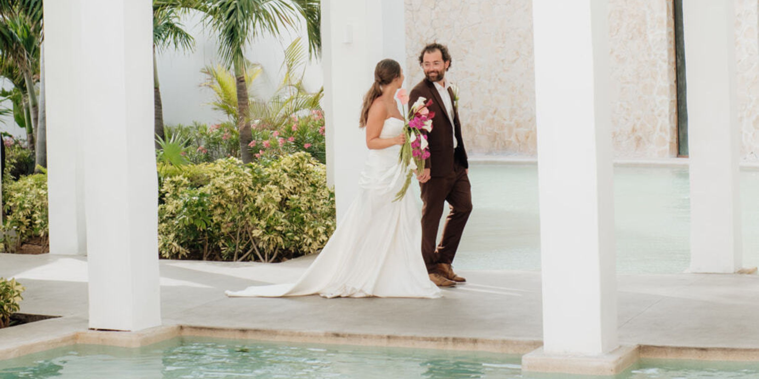 Ethan and Erica stroll by a pool at their destination wedding, framed by lush greenery and white pillars.