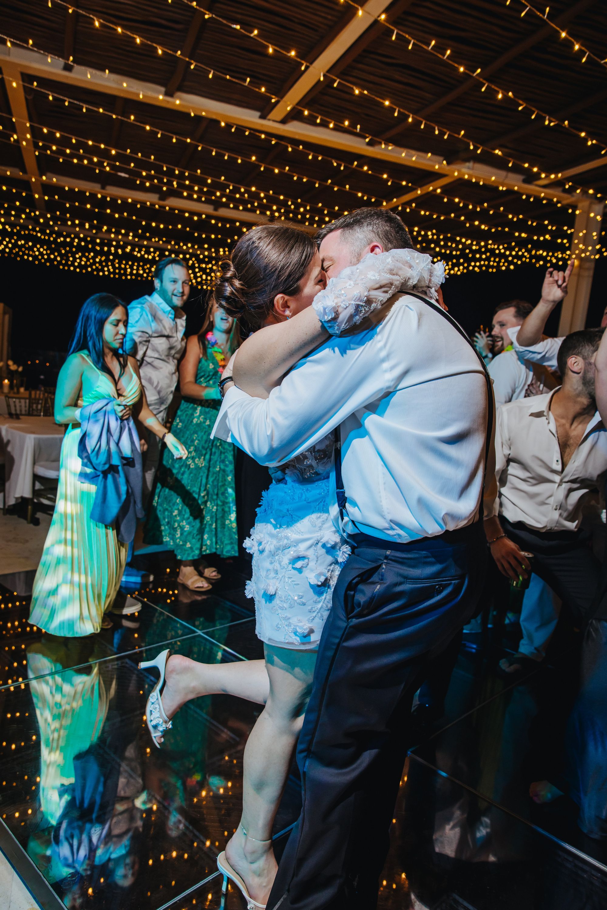 Kathryn and Mike share a kiss on the wedding dance floor under string lights, guests celebrating around them.