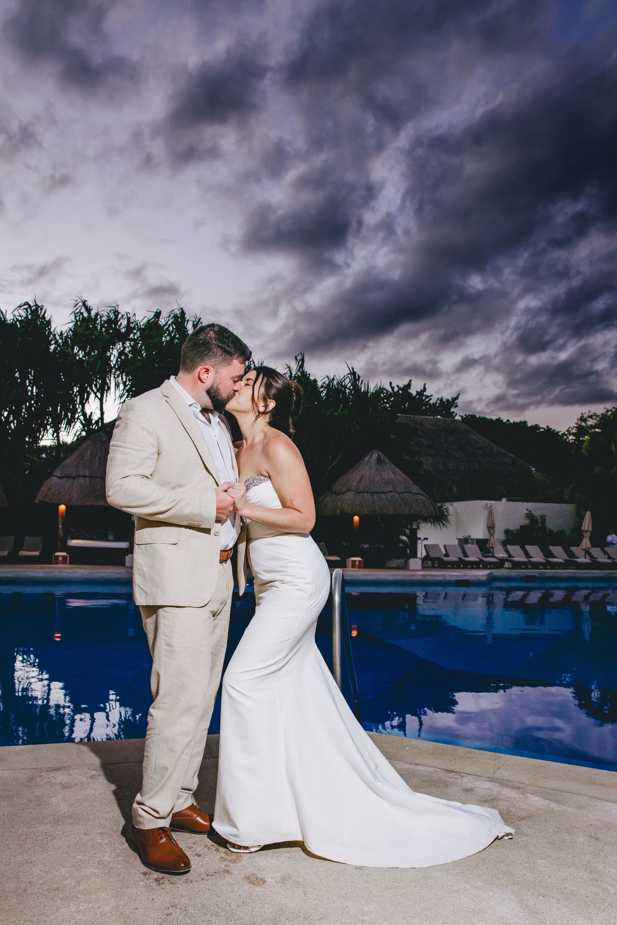 Kathryn and Mike kiss by the pool at dusk beneath dramatic clouds during their destination wedding.