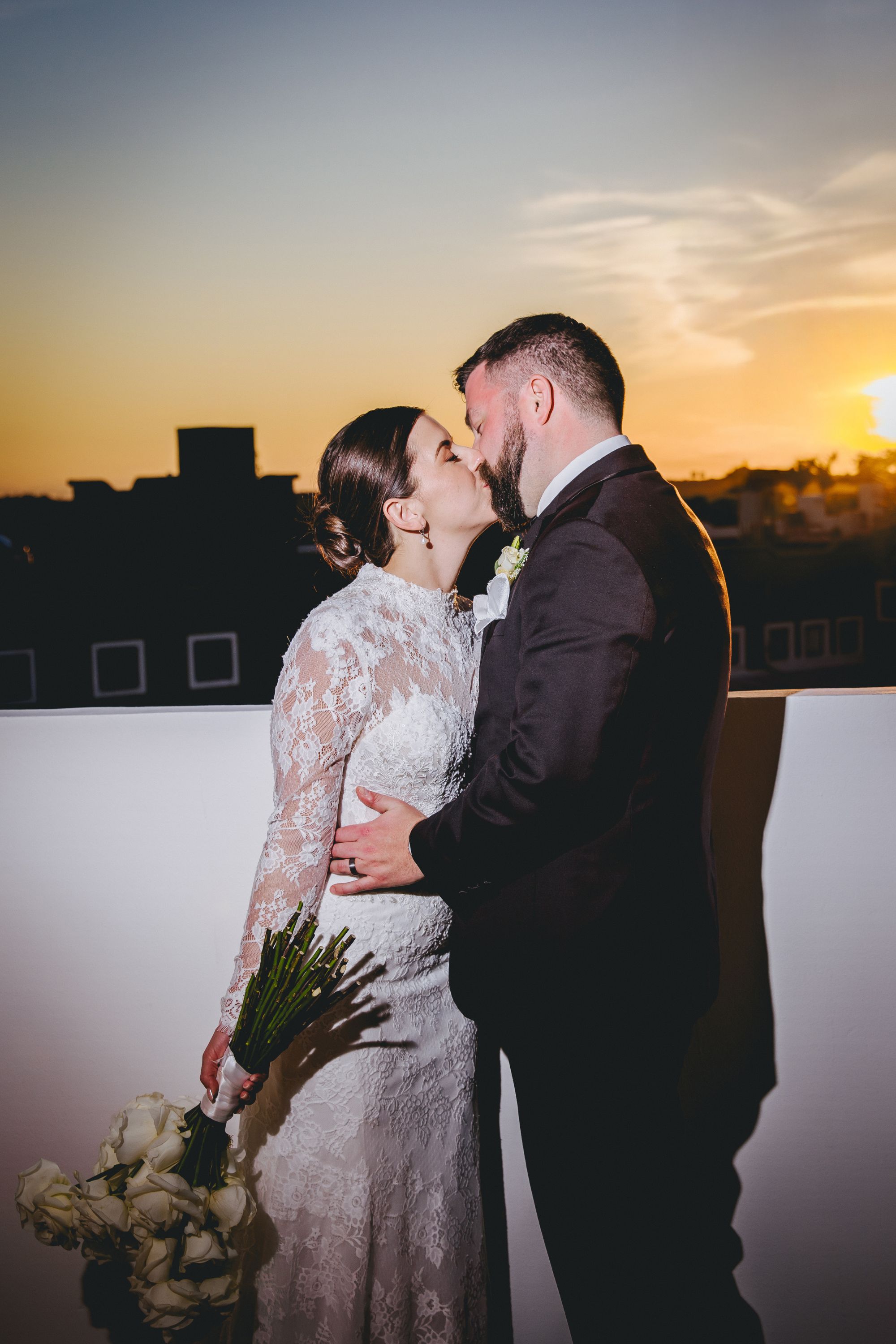 Kathryn and Mike share a sunset kiss on a rooftop at their destination wedding, white roses in hand.