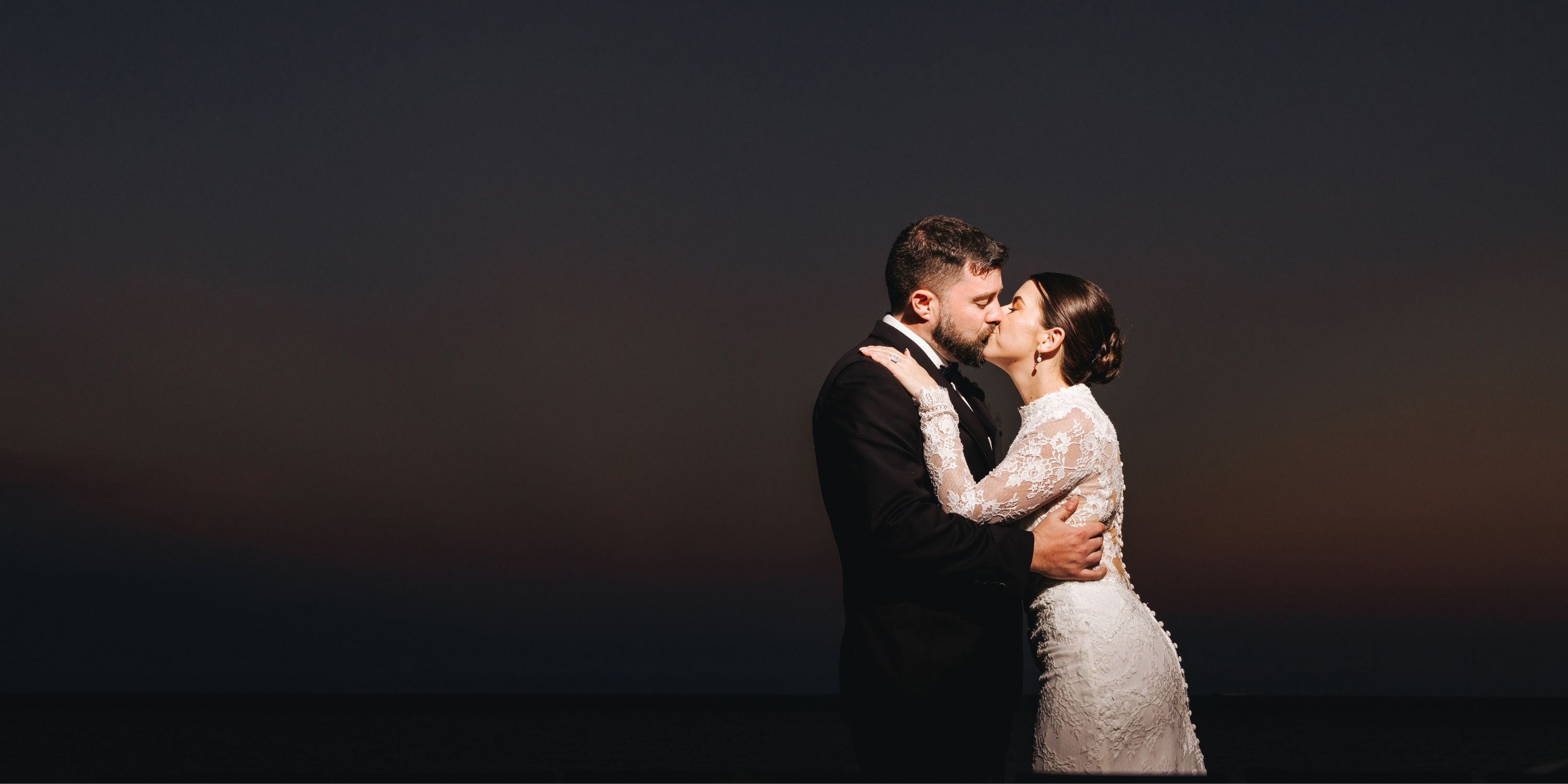 Kathryn and Mike share a romantic kiss at their destination wedding, embracing at dusk under a dark sky.