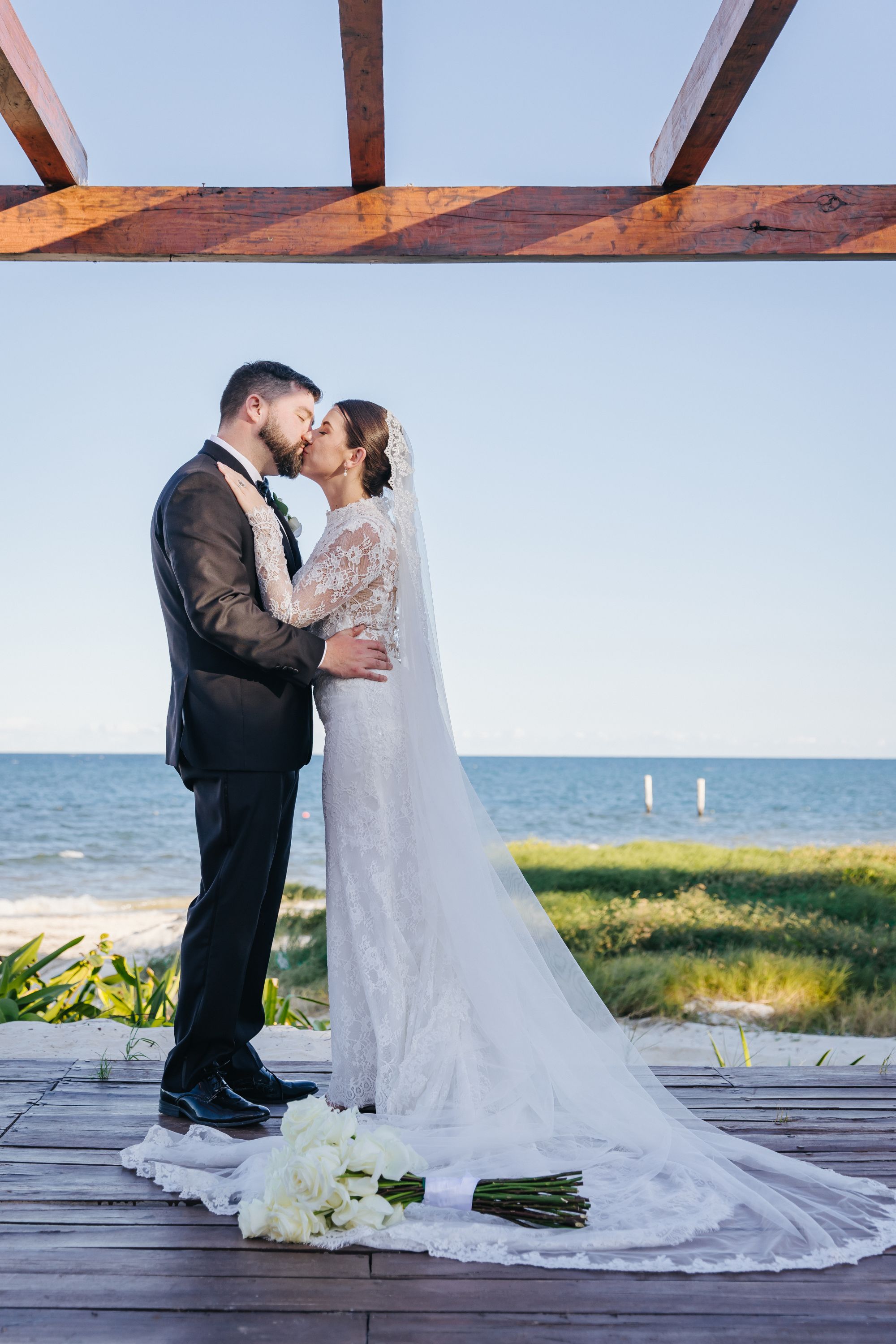 Kathryn and Mike share a kiss at their destination wedding under a pergola by the ocean, bouquet nearby.