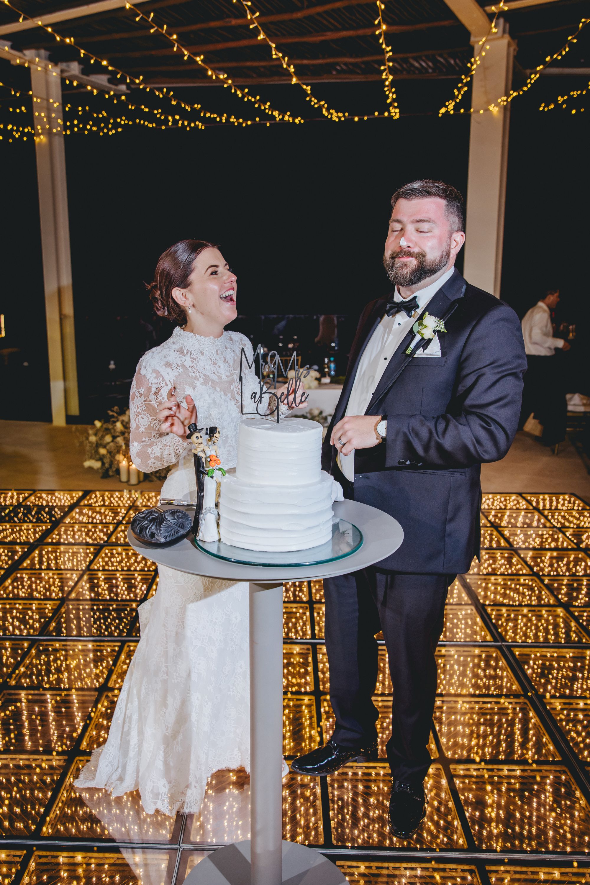 Kathryn and Mike pose by their wedding cake on a glowing dance floor beneath string lights at their wedding.