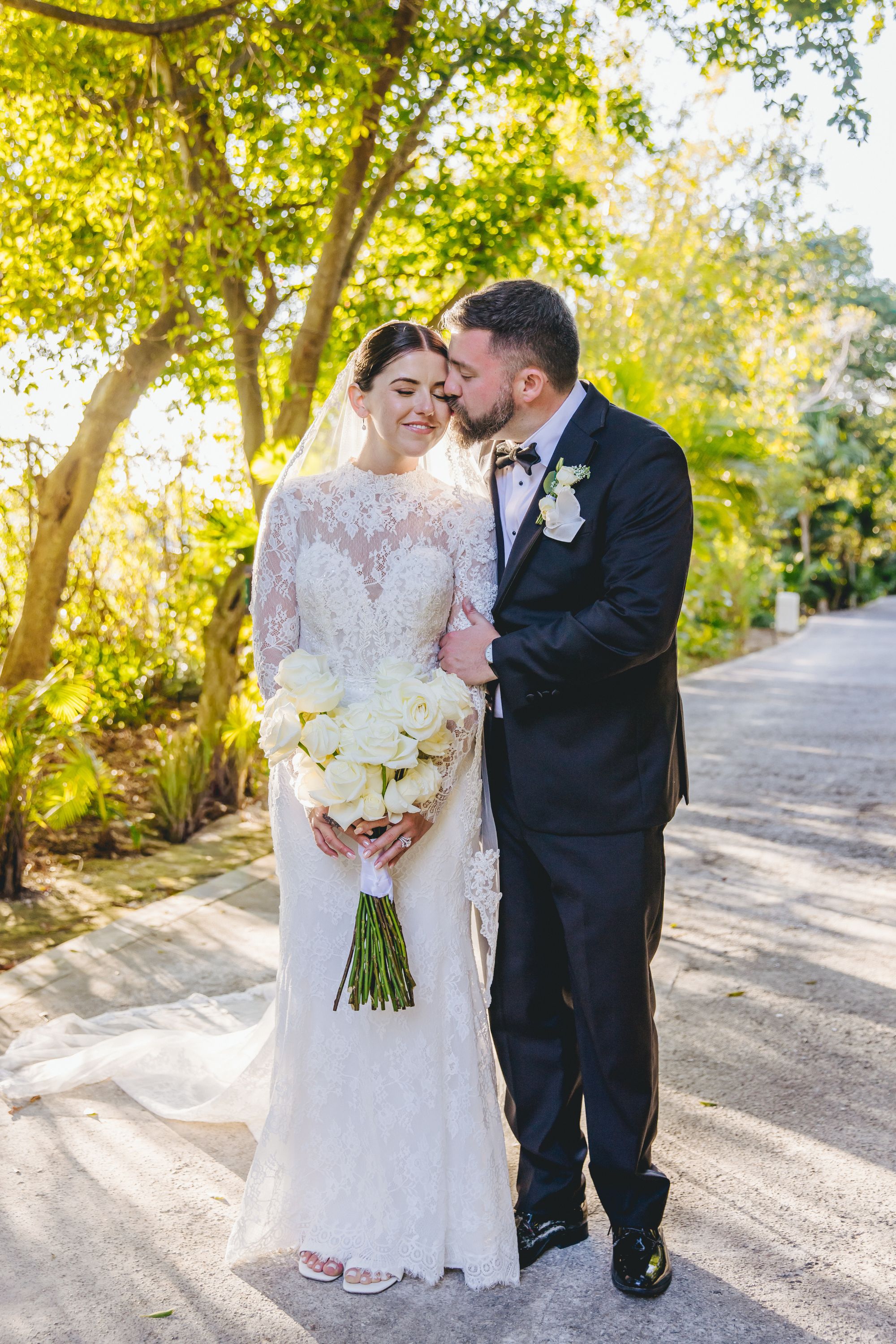 Bride in lace gown and groom in tux share a sweet forehead kiss at their outdoor destination wedding.