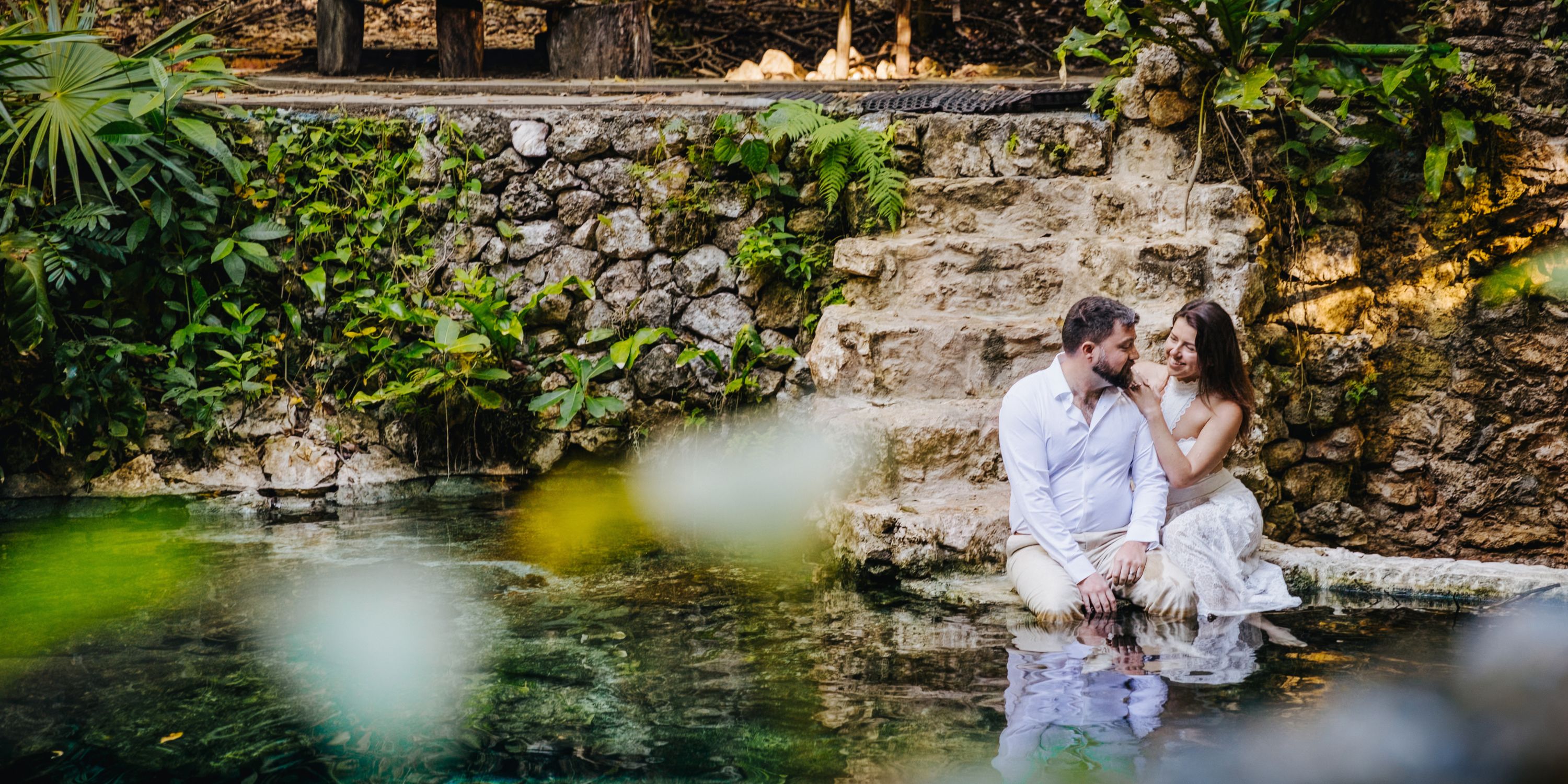 Kathryn and Mike (Copy) relax by a natural stone pool at their destination wedding, lush greenery around.