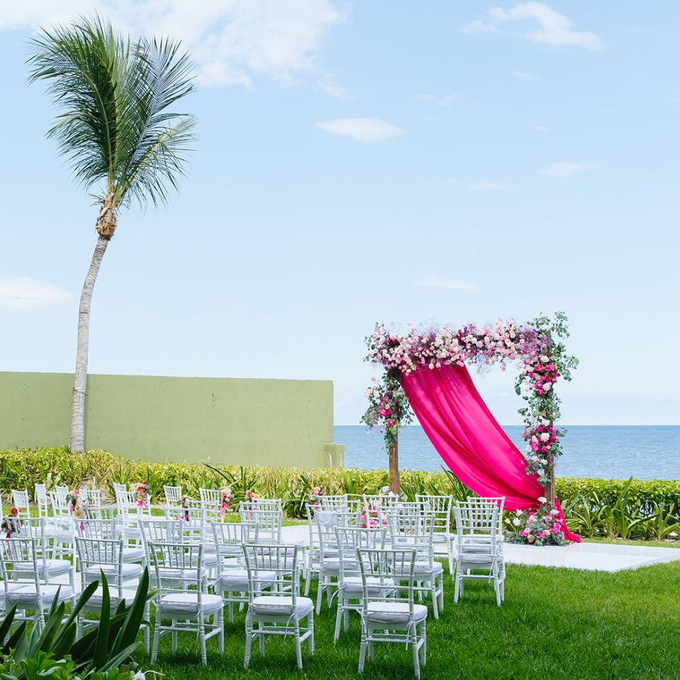 White chairs line up for a destination wedding, facing a floral arch with pink drapery by the ocean.