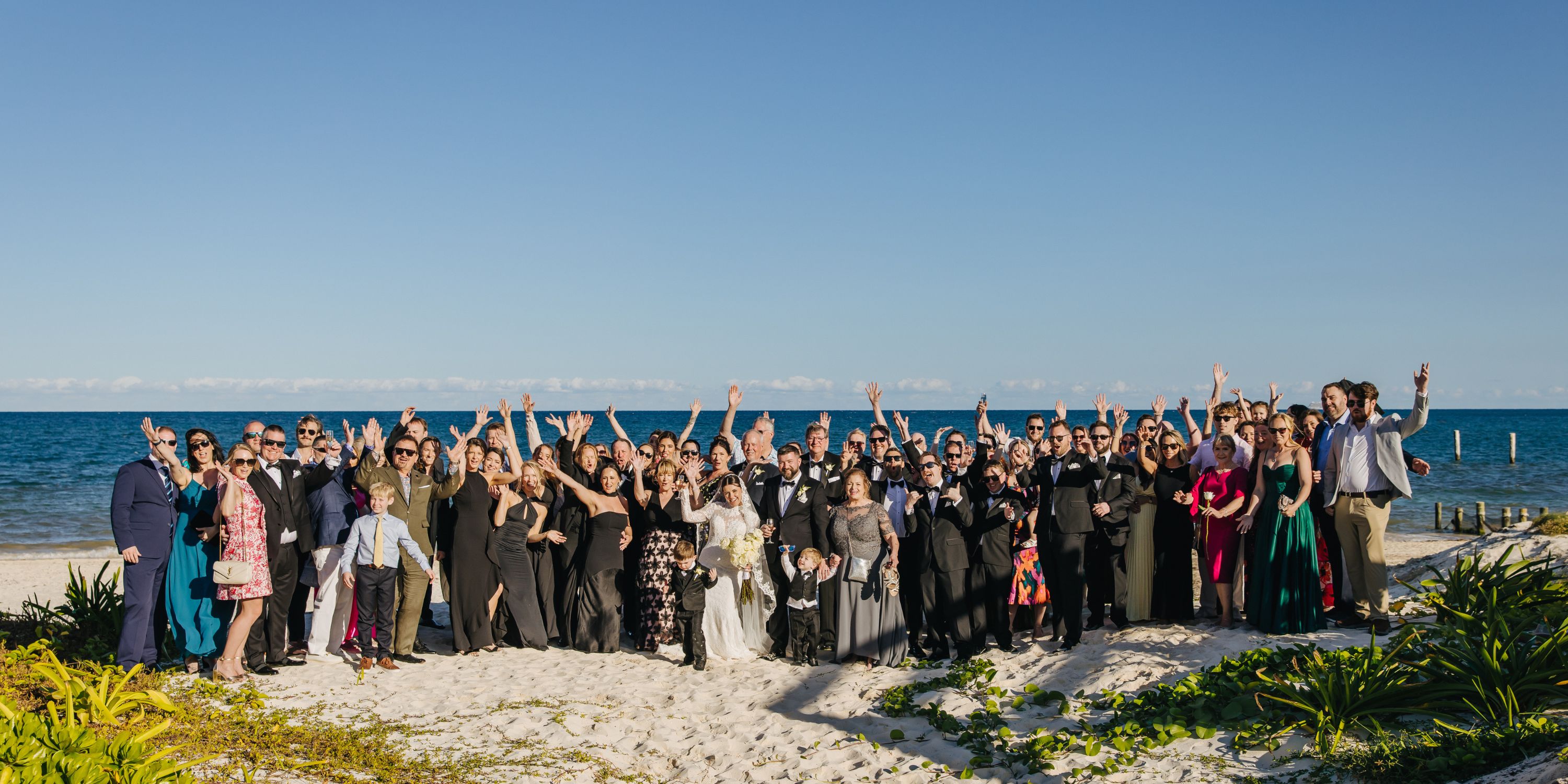 A large group poses on the beach at Kathryn and Mike's destination wedding, many with arms raised.