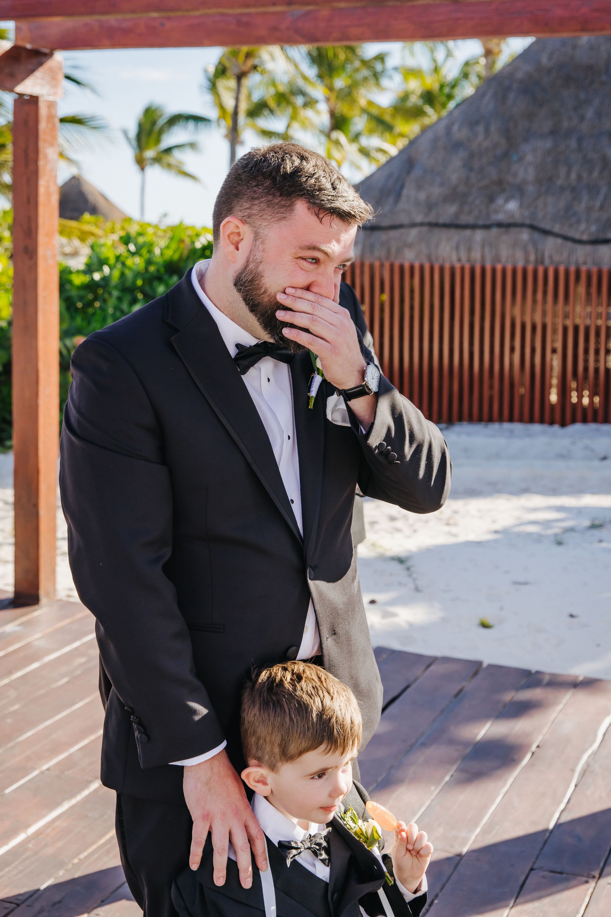 Man in tuxedo with young boy on a wooden deck at what appears to be a destination wedding outdoors.